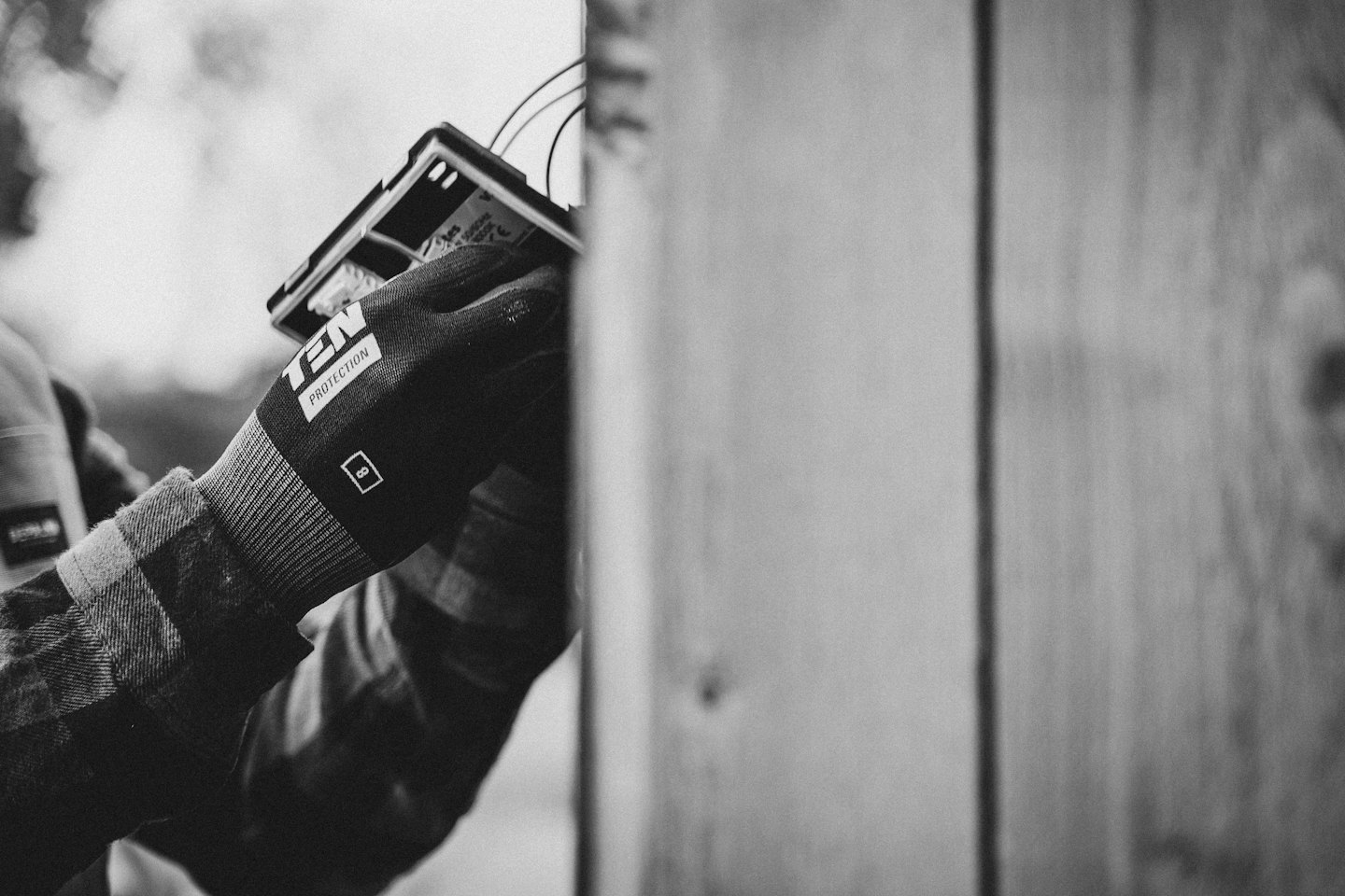 Gloved hands installing an electronic component near a wooden fence.